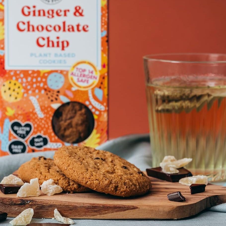 Vegan ginger and chocolate chip cookies on a wooden board with a box and glass of tea in the background.