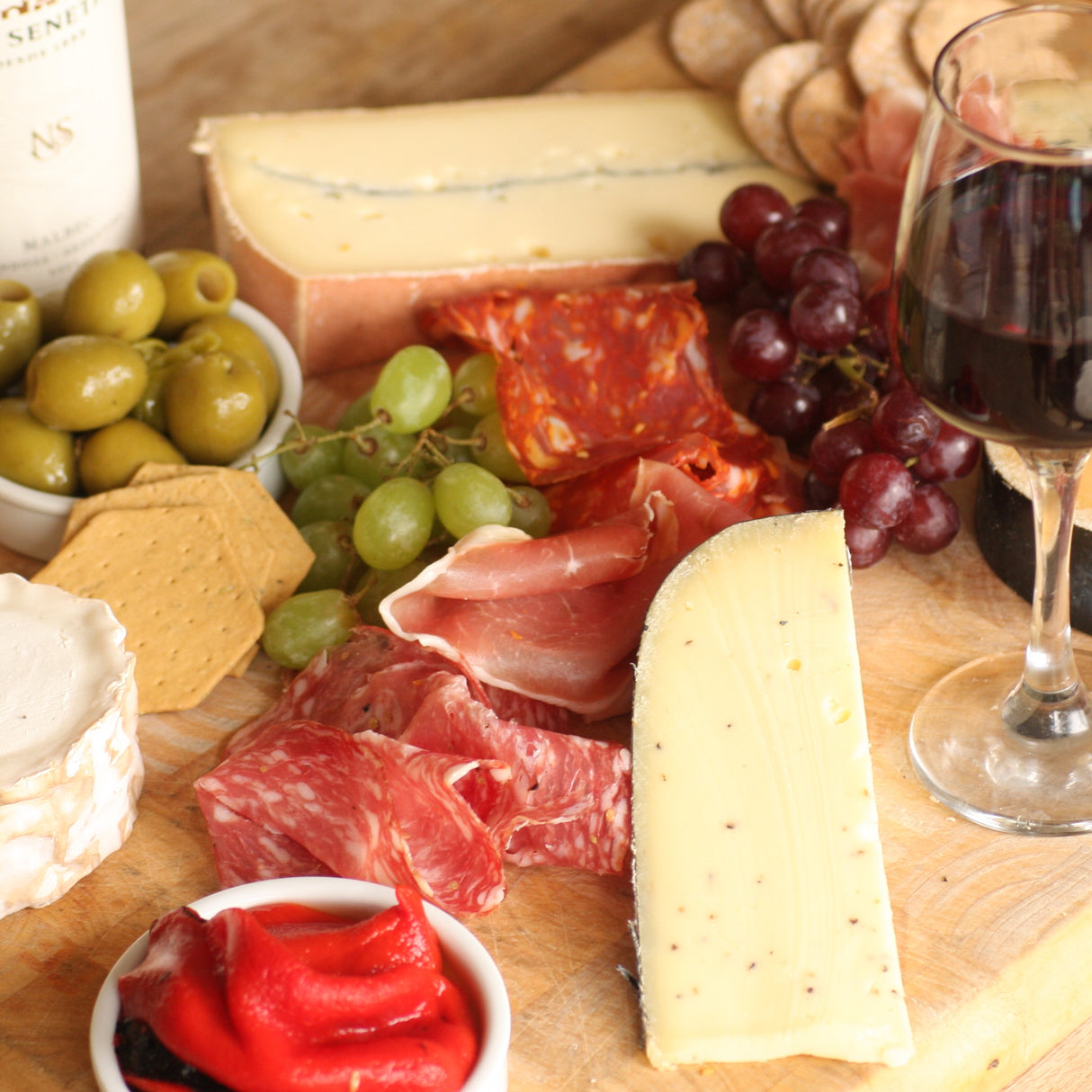 Assorted meats, cheeses, olives, and grapes on a wooden board with a glass of red wine.