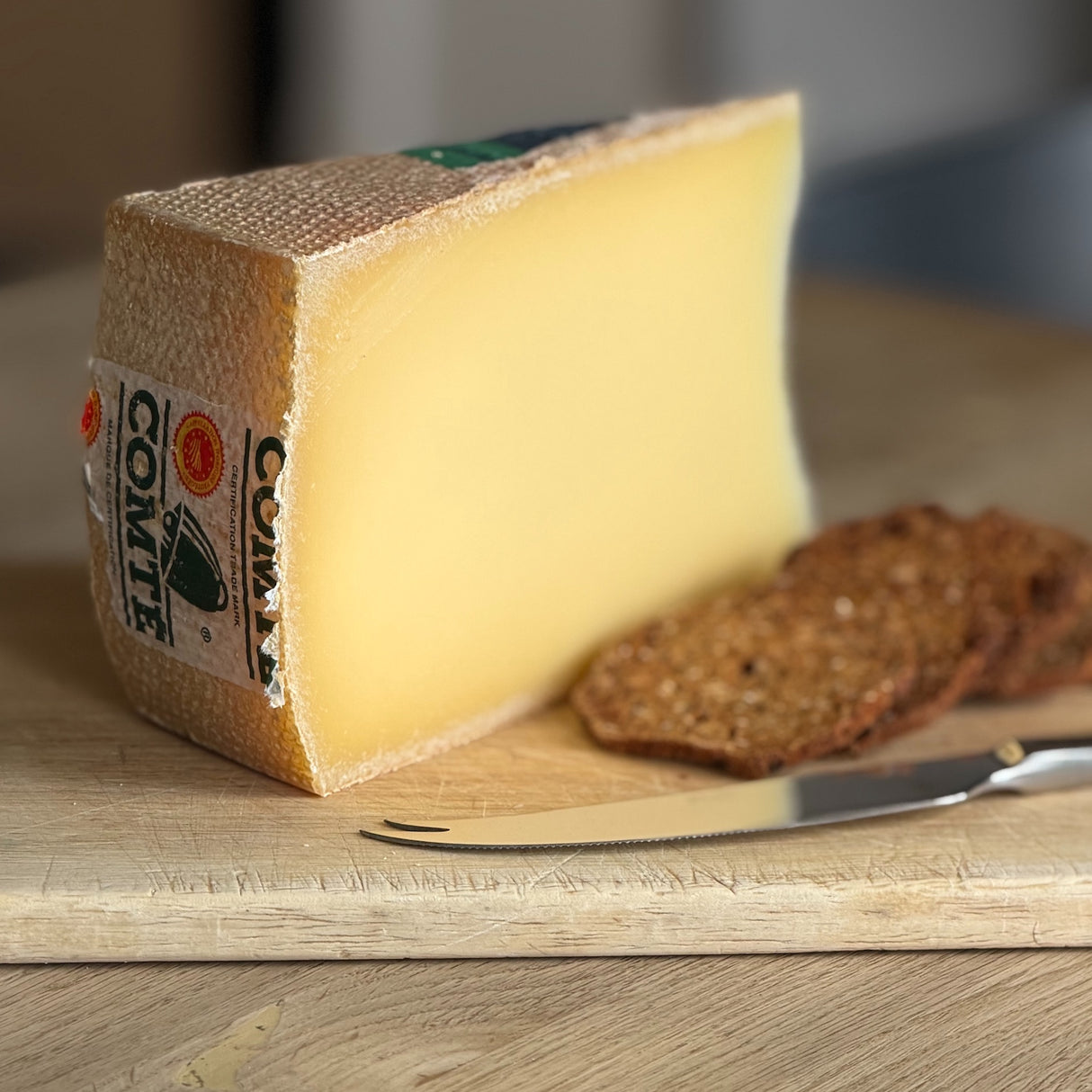 A wedge of Comte cheese on a wooden cutting board with a knife and two slices of rustic bread.