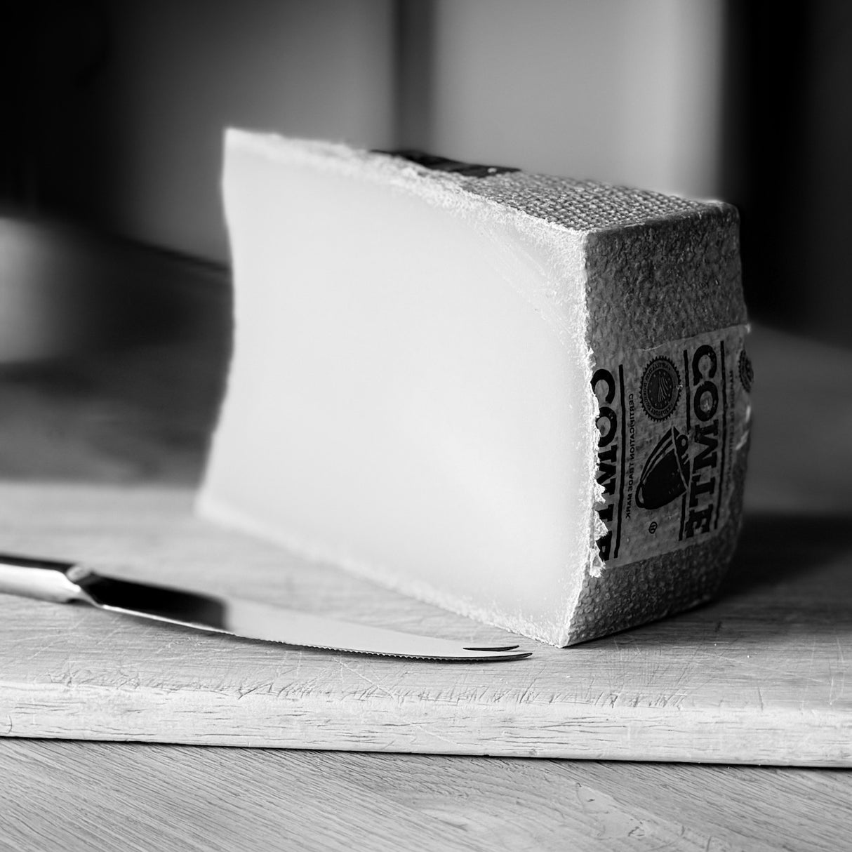 A wedge of Comte cheese on a wooden cutting board with a knife, shown in a black and white photograph.