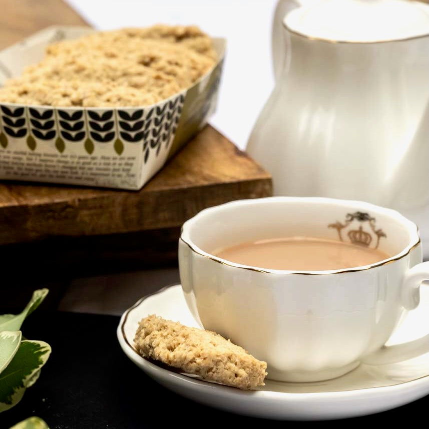 White teacup with tea and an oat flip biscuit on a saucer, next to a white teapot and a tray of oat flips.