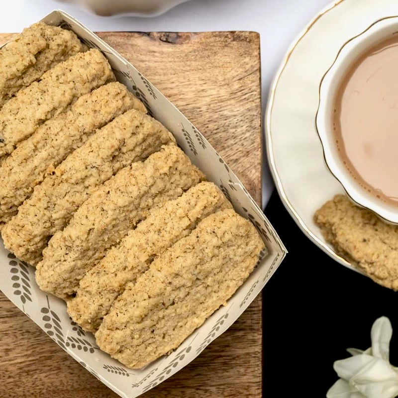 A tray of golden-brown, oat flip biscuits on a wooden board, with a cup of tea on a saucer.
