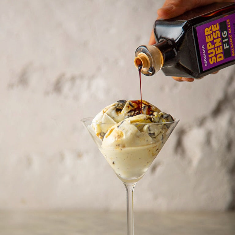A hand holds a bottle of fig balsamic glaze, drizzling it over a glass of ice cream