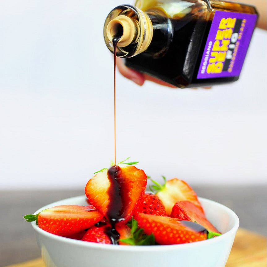 A hand holds a bottle of fig balsamic glaze over a white bowl filled with sliced strawberries, pouring a stream of the syrup over the fruit.