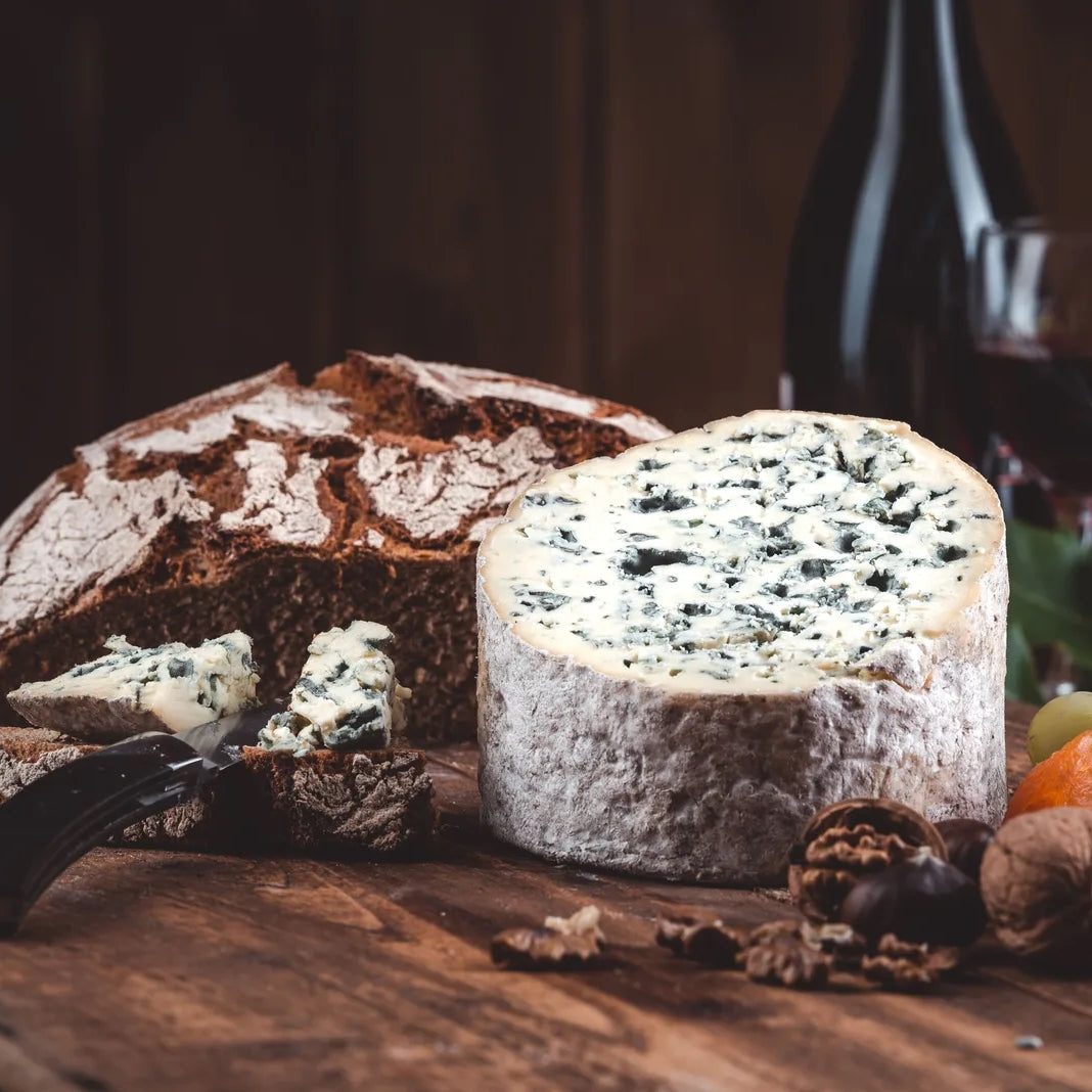 A round wheel of blue cheese stands on a wooden surface beside a loaf of rustic bread, with a knife, walnuts, and a wine bottle and glass blurred in the background.