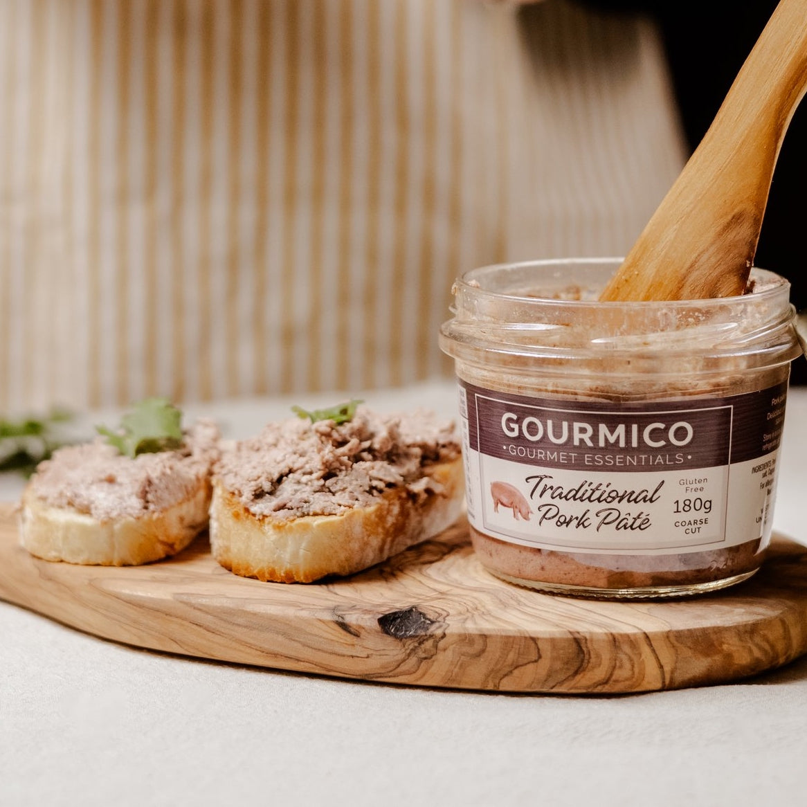 A jar of Gourmico Traditional Pork Pâté sits on a wooden serving board with two slices of bread topped with the pâté and a wooden spoon resting inside the jar.