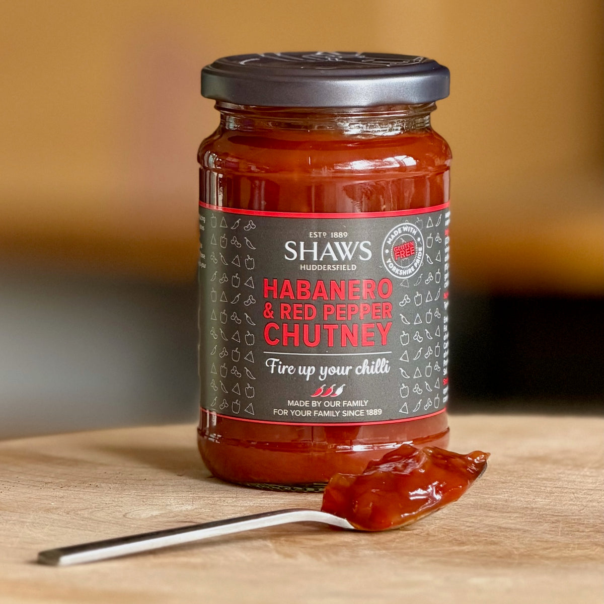 A glass jar of Shaw's Habanero & Red Pepper Chutney with a black lid and dark label, set on a wooden surface with a spoonful of the red chutney in front.
