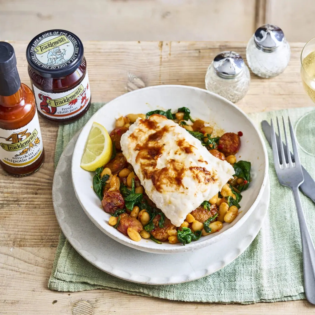 White plate with grilled fish over a bed of beans and greens with Tracklements Fresh Chilli Jam, on a wooden table.