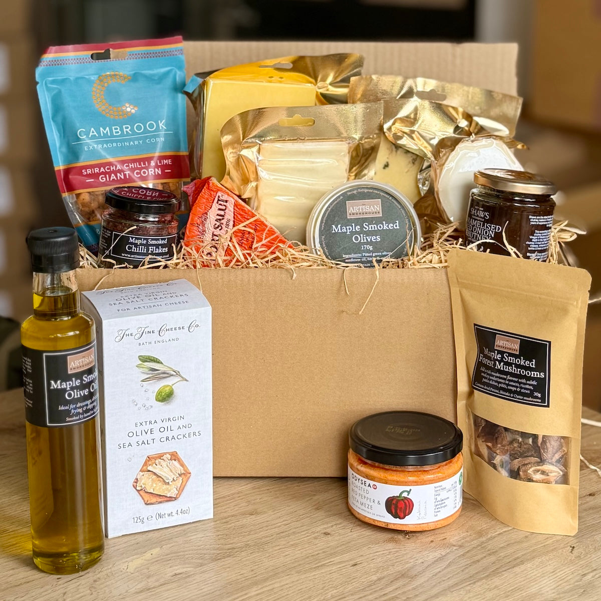 A gourmet gift hamper filled with assorted food items, including a bottle of maple smoked olive oil, a box of sea salt crackers, maple smoked olives, mushrooms, and other packaged snacks, arranged on a wooden surface with a cardboard box behind them.