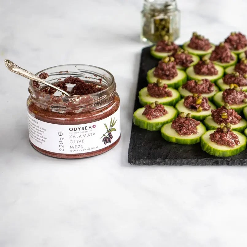 Jar of Odyssea Kalamata Olive Meze with a spoon and cucumber bites on a slate board.