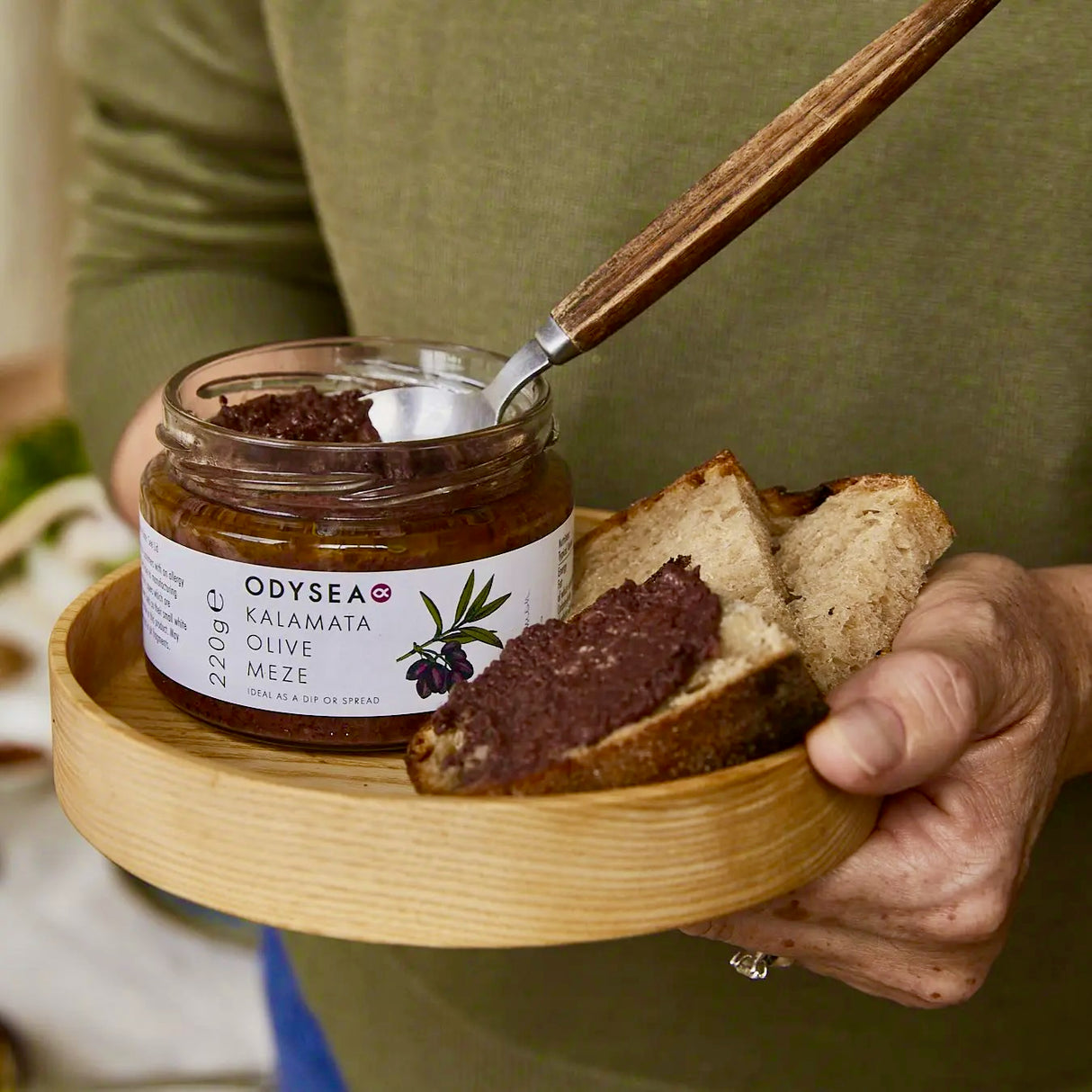 Person holding a jar of Odyssea Kalamata Olive Meze with bread on a wooden board.