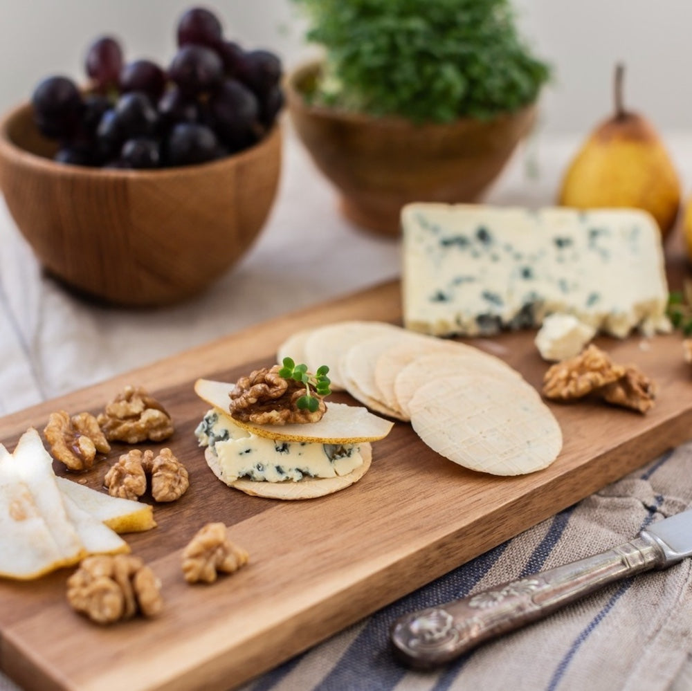 Wooden platter with Stilton cheese, gluten free crackers, and nuts on a table with bowls of grapes and pears in the background.