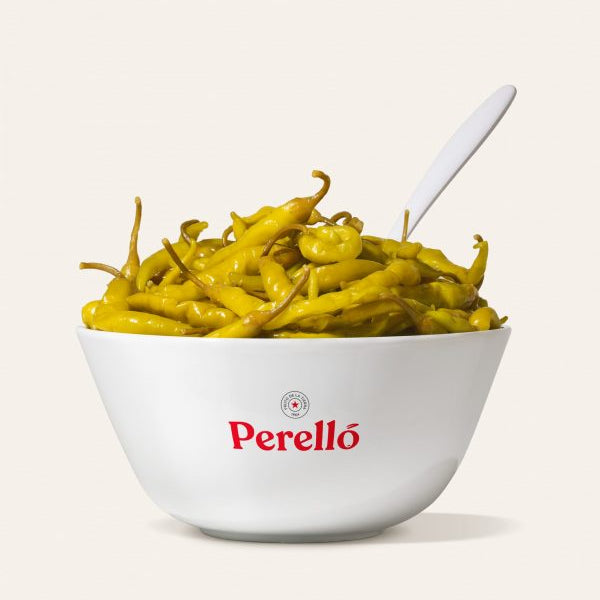 Bowl of pickled Guindilla peppers with a white spoon on a light background, featuring the Perello brand.