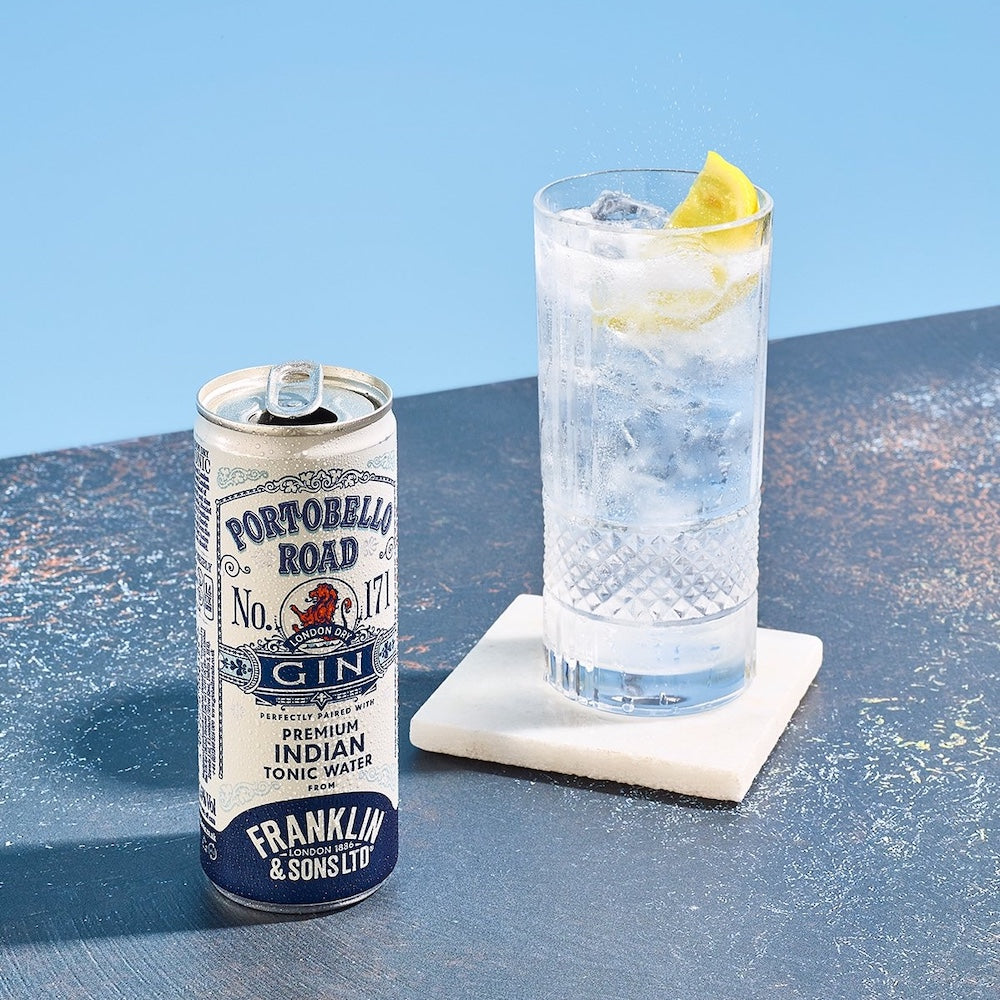 Can of Portobello Road Gin and Franklin & Sons tonic water with a glass of gin and tonic on a blue surface.