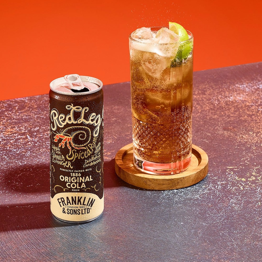 Red Leg Cola can and glass of rum & cola cocktail with ice and lime on a red background