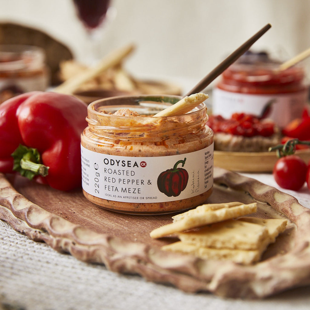 An open jar of Odysea Roasted Red Pepper & Feta Meze spread sits on a wooden board with crackers, red bell peppers, and tomatoes in a styled food setting.