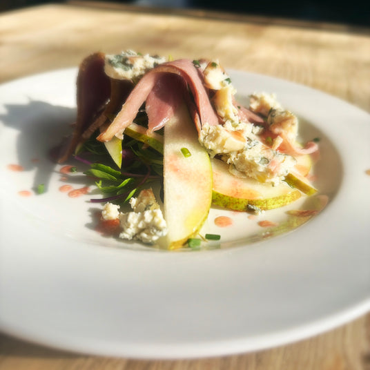 A plated gourmet salad featuring sliced pears, crumbled blue cheese, green herbs, and slices of smoked duck on a white plate, set on a light wooden surface.