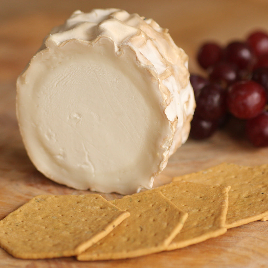 A round wheel of creamy goats cheese with a textured rind, shown on a wooden surface with crackers in front and red grapes in the background.