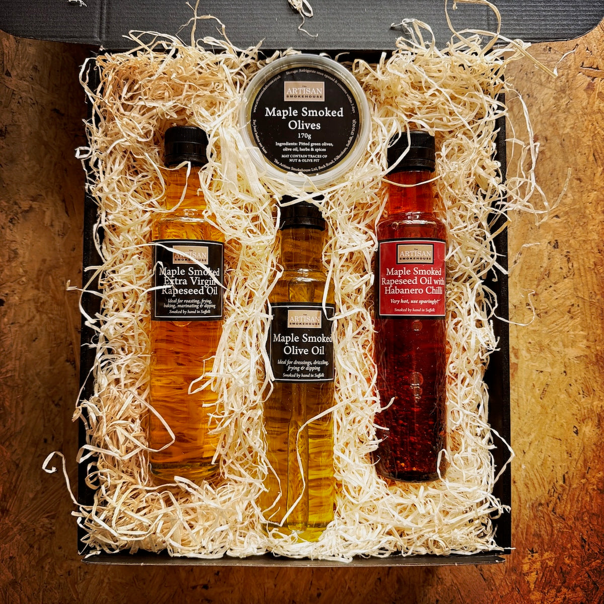 A gift set of maple-smoked oils and smoked olives arranged in a black box filled with shredded wood on a wooden surface.