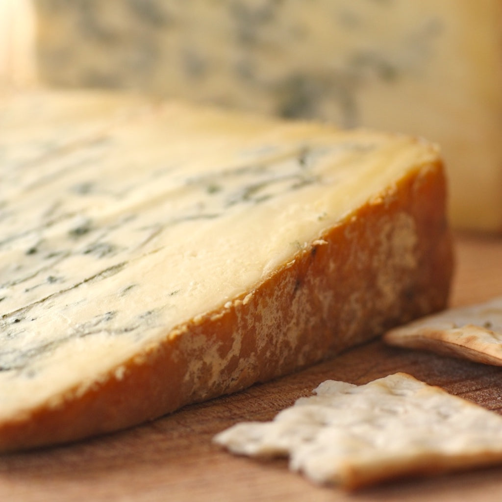 A close-up of a wedge of smoked Stilton cheese on a wooden surface with crackers in the foreground and a blurred cheese block in the background.