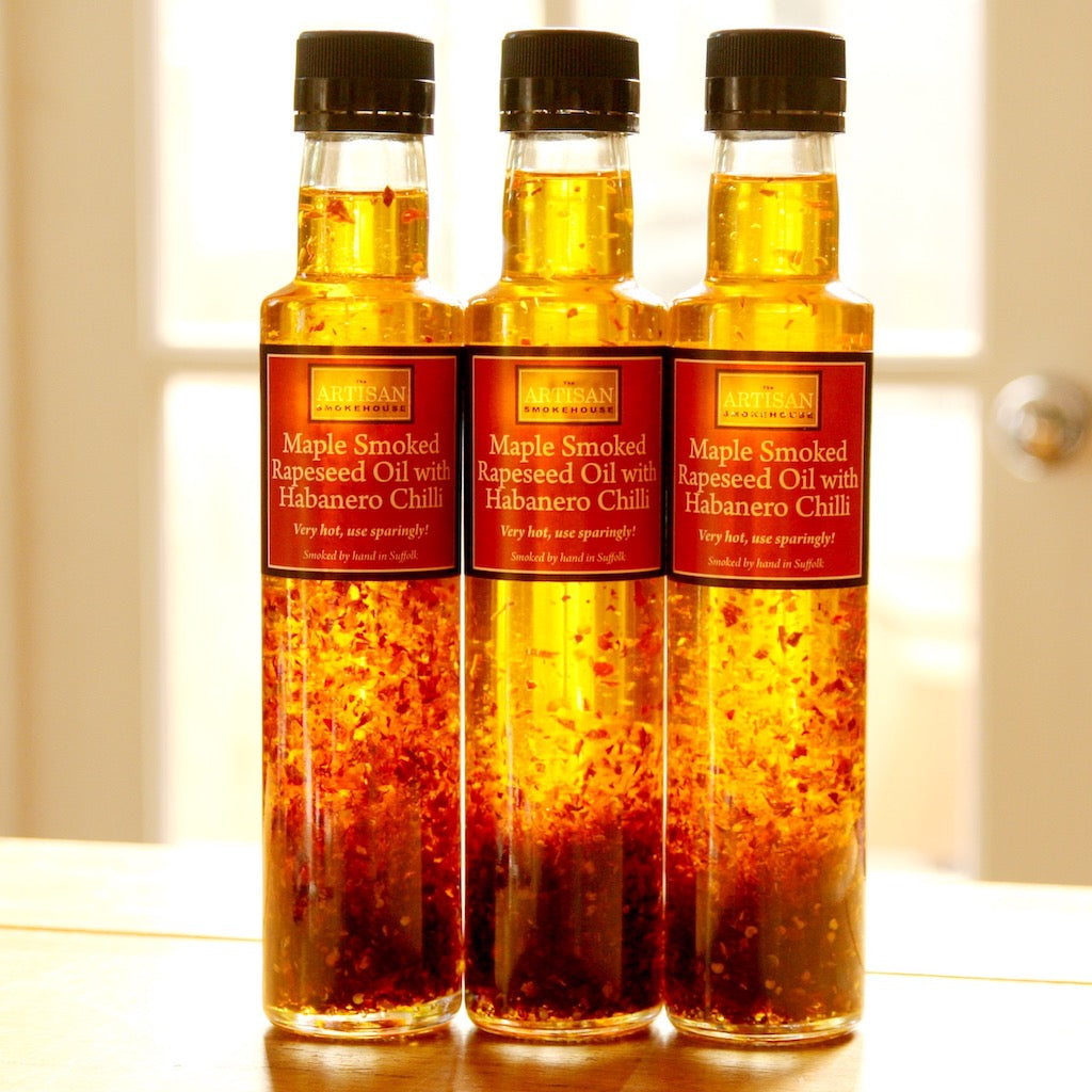 Three glass bottles of Maple Smoked Rapeseed Oil with Habanero Chili, filled with golden oil and red chili flakes, standing on a wooden surface.