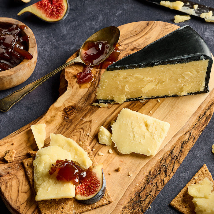 A styled cheese board featuring a wedge of Snowdonia Black Bomber, crackers topped with cheese and fig jam, a spoon of jam, and a small bowl of figs on a dark slate background.