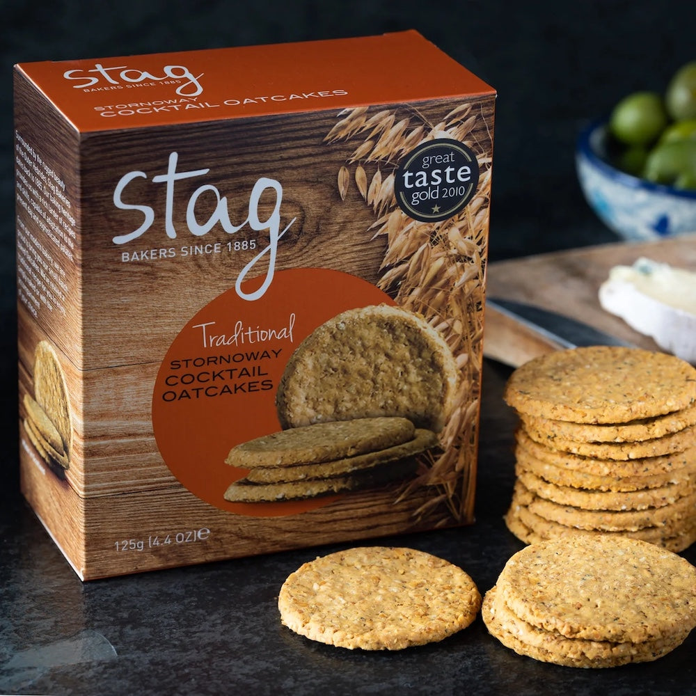 A box of Stag Traditional Stornoway Cocktail Oatcakes with several round oatcake slices stacked and placed in front on a dark tabletop.