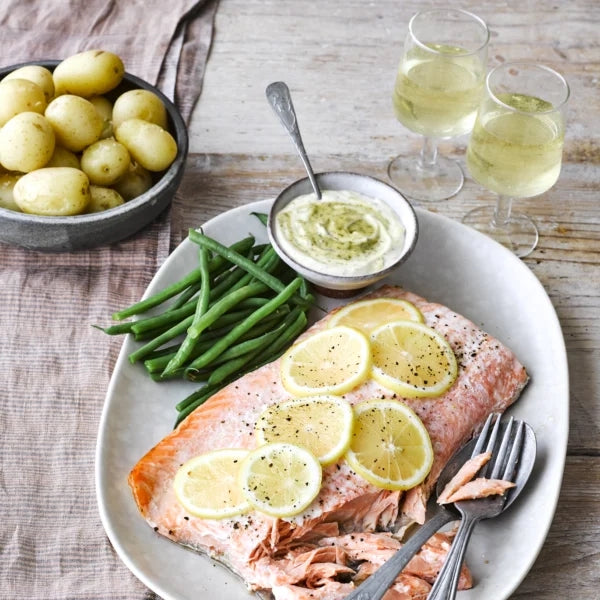Plated salmon with lemon slices, green beans, and potatoes on a wooden table with wine glasses and Tracklements
dill mustard sauce served in a bowl.