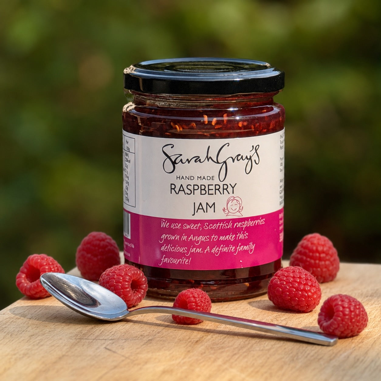 A jar of Sarah Grey's Hand Made Raspberry Jam sits on a wooden board surrounded by fresh raspberries and a silver spoon.