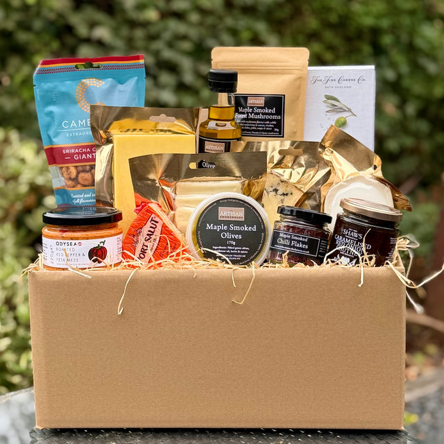 A gourmet gift box filled with various food items, including jars of chutney, olives, cheeses, a bottle of olive oil, and packaged snacks, arranged in a brown cardboard box with shredded paper on a table outdoors.