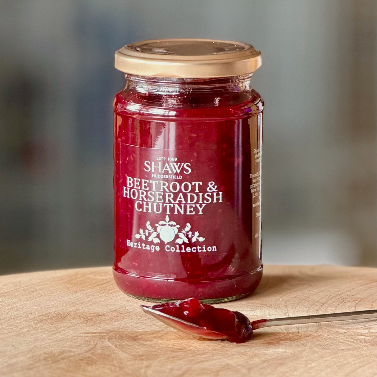 A glass jar of Shaws Beetroot & Horseradish Chutney sits on a wooden cutting board with a spoonful of the deep red chutney in front. 