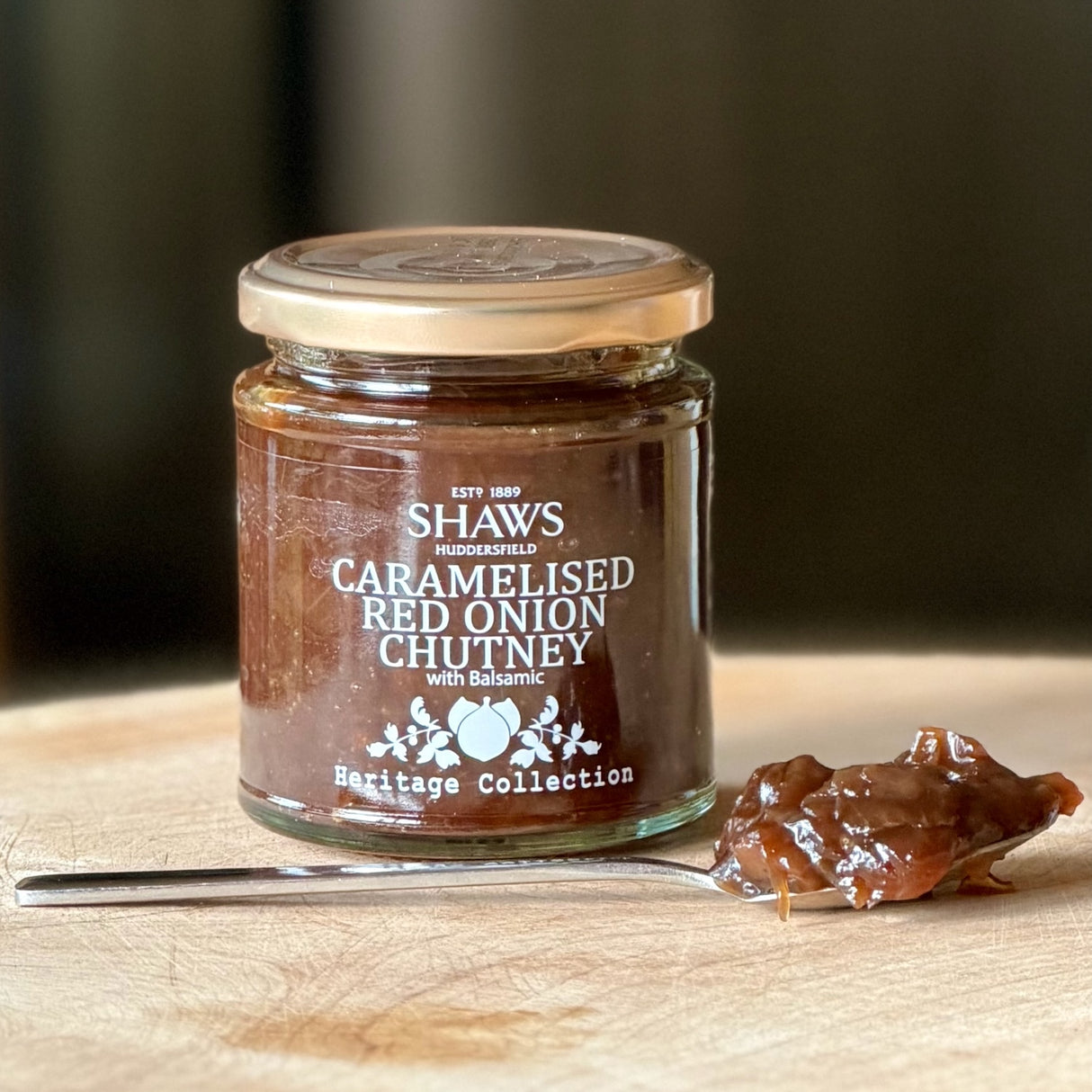 A glass jar of Shaws Caramelised Red Onion Chutney with Balsamic on a wooden cutting board, with a metal spoon holding some of the thick brown chutney in front.