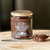 A glass jar of Shaws Caramelised Red Onion Chutney with Balsamic on a wooden cutting board, with a metal spoon holding some of the thick brown chutney in front.