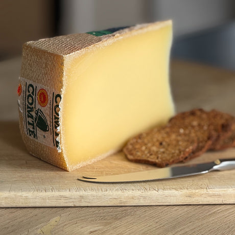 A wedge of Comte cheese on a wooden cutting board with a knife and two slices of rustic bread.