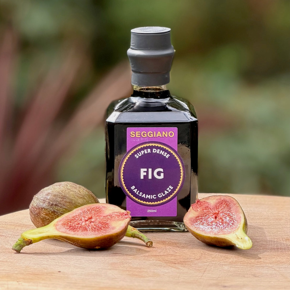 A bottle of Seggiano Super Dense Fig Balsamic Glaze sits on a wooden surface with fresh figs, including one whole fig and two halved figs.