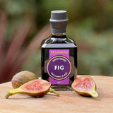A bottle of Seggiano Super Dense Fig Balsamic Glaze sits on a wooden surface with fresh figs, including one whole fig and two halved figs.