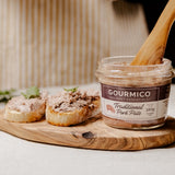 A jar of Gourmico Traditional Pork Pâté sits on a wooden serving board with two slices of bread topped with the pâté and a wooden spoon resting inside the jar.