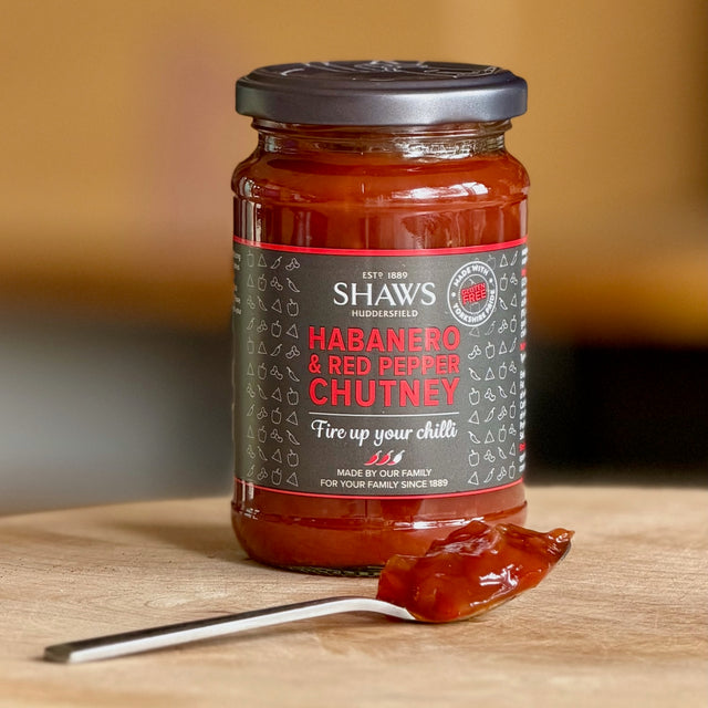 A glass jar of Shaw's Habanero & Red Pepper Chutney, set on a wooden surface with a spoonful of the red chutney in front.
