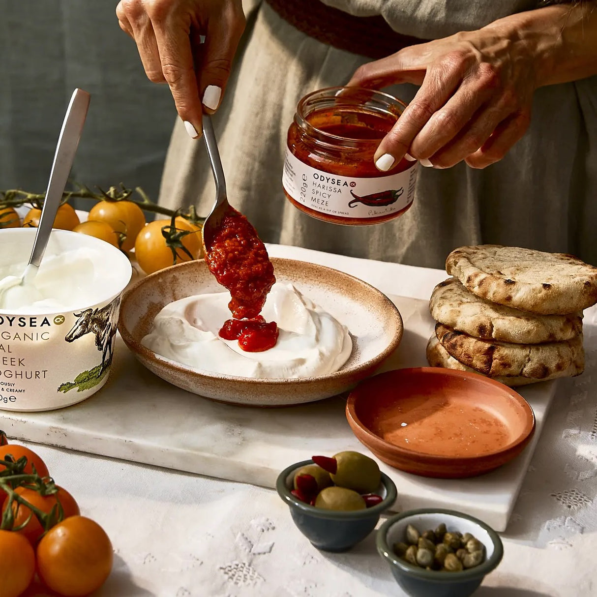 Person spooning harissa Meze on yogurt, surrounded by bread and vegetables.