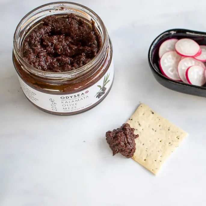 Jar of olive tapenade with a spreadable portion on a cracker, set against a white background.