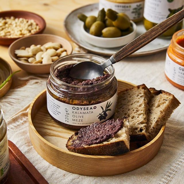 Jar of Odyssey Kalamata Olive Meze spread on a wooden plate with bread slices.