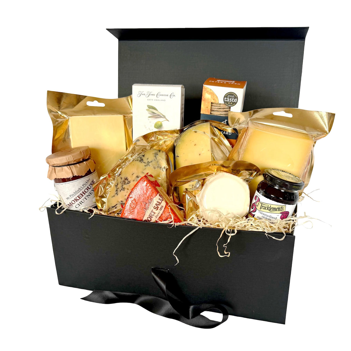 A luxury cheese gift hamper set in a black box with a black ribbon, filled with various cheeses, crackers, and preserves on a white background.