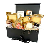 A luxury cheese gift hamper set in a black box with a black ribbon, filled with various cheeses, crackers, and preserves on a white background.