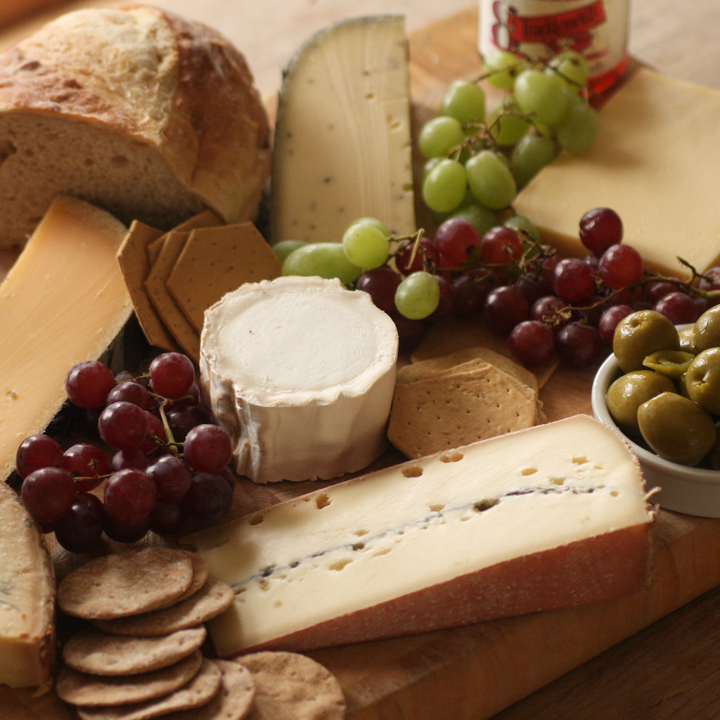 A piece of Morbier cheese on cheese board with other cheeses, crackers, bread, grapes, chutney and smoked olives