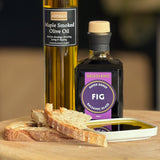 A bottle of Fig Balsamic Glaze and a bottle of Maple Smoked Olive Oil on a wooden board with slices of bread and a small dish of the dark balsamic oil.