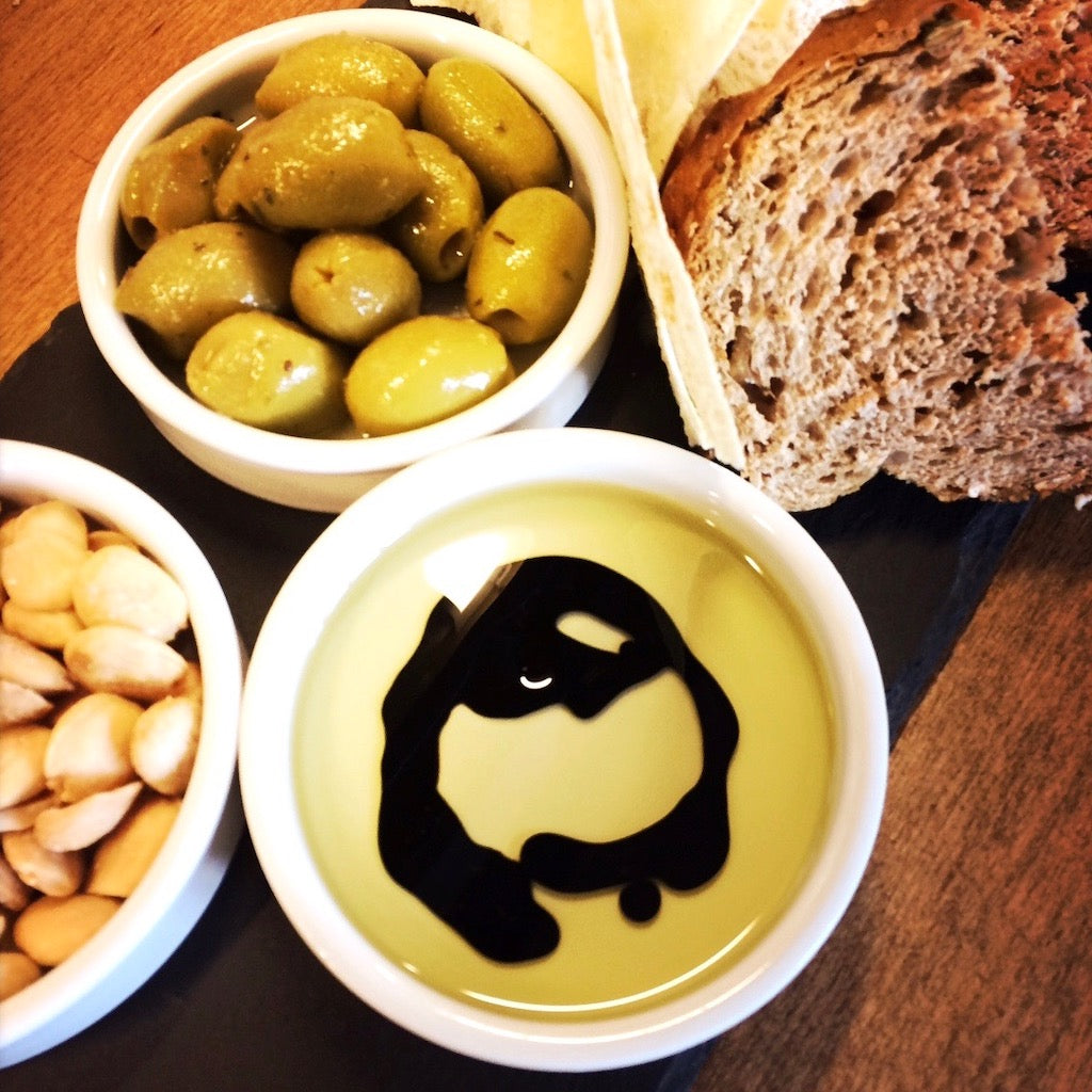 A ramekin with The Artisan Smokehouse's smoked olive oil & balsamic vinegar with dipping bread, smoked olives and nuts on platter