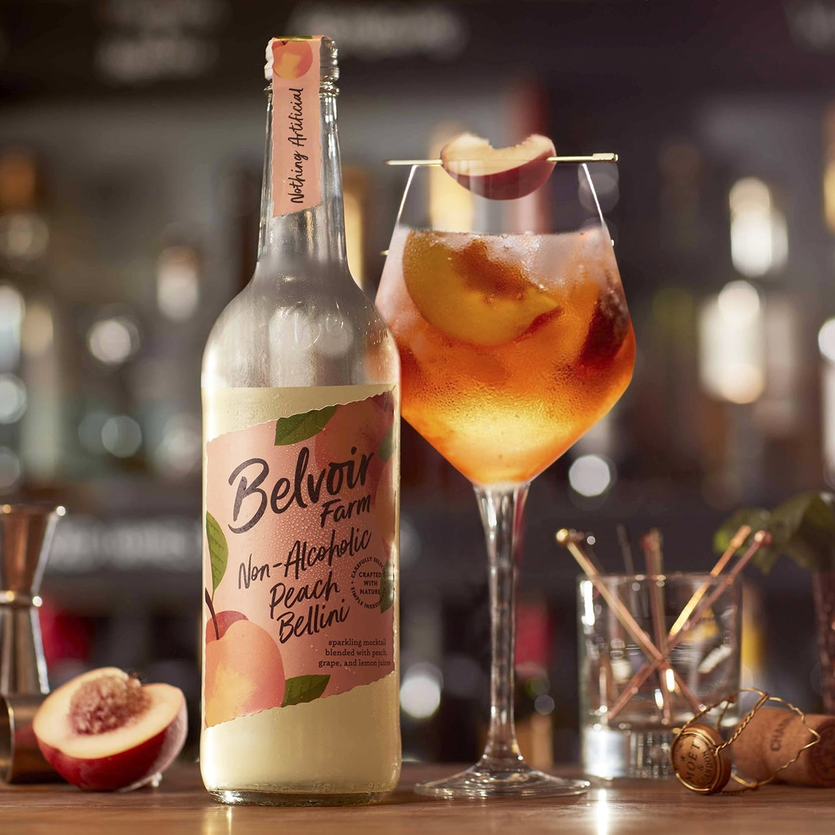 A bottle of Belvôir Farm Non-Alcoholic Peach Bellini cocktail mix stands on a bar counter beside a prepared peach-colored drink in a coupe glass.