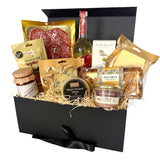 A gourmet picnic hamper filled with assorted cheeses, cured meats, crackers, and a bottle of drink, arranged inside an open black gift box.