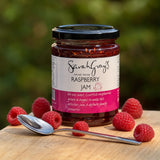 A jar of Sarah Grey's Hand Made Raspberry Jam sits on a wooden board surrounded by fresh raspberries and a silver spoon.