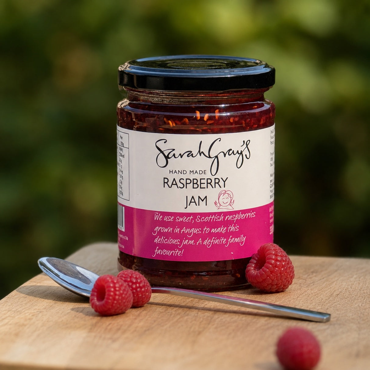 A jar of Sarah Grey's Hand Made Raspberry Jam sits on a wooden board with fresh raspberries and a spoon.
